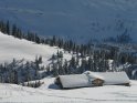 Blick über die von Ski-Spuren durchzogene Schneelandschaft auf ein schneebedecktes Haus mit eingeschneiten Tannenbäumen im Hintergrund