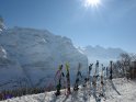 Eine Reihe von in den Schnee gesteckten Skiern mit den schneebedeckten Alpen im Hintergrund.