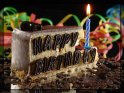 Stück Kuchen mit einer Kerze und dem Schriftzug 
 Happy Birthday aus Schokolade auf die Seite 
 geschrieben. Im Hintergrund sind Luftschlangen zu sehen.