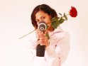 Eine junge Frau hält eine rote Rose im Mund und präsentiert dabei einen Akkuschrauber.