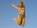 Komplett mit Sand bedeckte Frau springt vor blauem Himmel in die Luft.