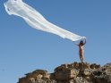 Eine Frau steht im Bikini an einer Felsenklippe und lässt ein riesiges weißes Stück Stoff vor blauem Himmel fliegen.