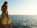 Eine Frau mit Hawaiikette und Bastrock steht auf einem Felsen am Meer.