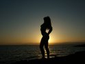 Schlanke Frau posiert bei Sonnenuntergang am Meer vor der Sonne. Somit ist lediglich ihre Silhouette zu erkennen.