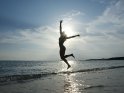 Eine Frau im Bikini springt im flachen Wasser in die Luft. Da die Sonne direkt hinter ihr steht, lässt sich nur ihre Silhouette erkennen.