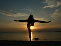 Silhouette einer auf einem Bein stehenden und die Arme zur Seite streckenden Frau bei Sonnenuntergang mit dem Meer im Hintergrund.