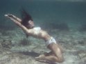 Eine Junge Frau im weißen Bikini kniet unterwasser auf dem Meeresboden.