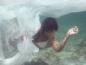 Eine Frau im weißen Bikini taucht unter einem weißen halb transparenten Tuch hindurch. Um Hintergrund ist der Meeresboden zu erkennen.