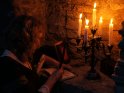Frau in Gewandung schreibt mit einer Feder in einem Buch. Beleuchtet wird die ganze Szene von einem Kerzenständer mit fünf brennenden Kerzen.