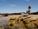 Eine Frau im Sommerkleid sitzt auf den Felsen am Strand. Dahinter ist der Strand zu erkennen.