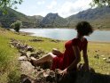 Eine Frau in einem roten Sommerkleid sitzt auf Steinen an einem Bergsee.