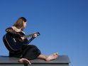 Junge Frau im schwarzen Kleid sitzt auf einem Dach vor blauem Himmel und spielt auf einer schwarzen Gitarre.