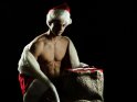 Sexy Foto von einem Weihnachtsmann, der mit einem vorne geöffneten Mantel neben dem mit Geschenken gefüllten Sack kniet. Durch die Pose ist sein muskulöser Oberkörper gut zu erkennen.