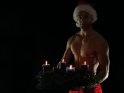 Sexy Weihnachtsmann mit freiem Oberkörper hält einen Adventskranz mit vier brennenden Kerzen.