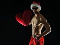 Foto eines muskulösen Weihnachtsmanns, der mit freiem Oberkörper eine Stoffherz in der Hand hält. Neben einer Weihnachtsmütze trägt er auch eine dunkle Sonnenbrille.