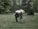 Familie spaziert durch den Regen. Dabei hat der Vater die beiden Kinder unter die Arme geklemmt während die Mutter mit Regenschirmen hinterher läuft.