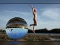 Foto von einer auf dem Boden liegenden Glaskugel, in der man eine junge Frau sieht, die im Bikini an einem See in die Luft springt.