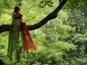 Junge Frau sitzt in einem Kleid, bestehend aus einem roten und einem grnen Seidentuch auf einem Baum.