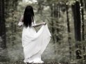 Eine Frau steht im weißen Brautkleid mit dem Rücken zur Kamera im Wald. Um sie herum sieht man dünne Nebelschwaden.