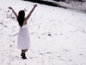 Eine Frau steht im weißen Sommerkleid auf einer verschneiten Wiese und streckt vor Freude die Arme in die Luft.