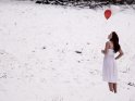 Eine junge Frau steht in einem weißen Sommerkleid im Schnee und hält dabei zwei herzförmige Heliumballons in der Hand.