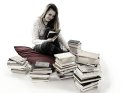 Eine junge Frau sitzt um geben von Bücherstapeln auf einem Kissen und liest in einem Buch.