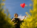 Eine Frau steht im schwarzen Kleid im Rapsfeld und hlt einen rote Heliumballon in Herzform in der Hand.